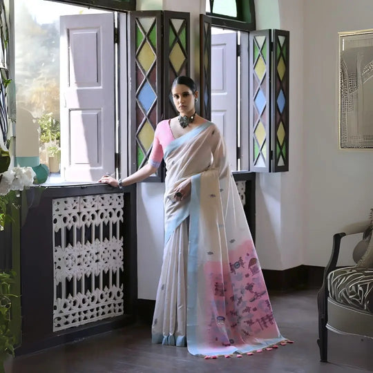 A woman models a graceful Off White Soft Muga Cotton Silk Saree. The saree has a light blue border, and the pallu features a wide pink panel decorated with traditional Warli-style figurative motifs in blue and purple. She wears a pink blouse and a dark oxidized choker necklace, posing indoors.