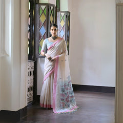A woman models a graceful Off White Soft Muga Cotton Silk Saree. The saree has a dark pink woven border, and the pallu features a wide sky blue panel decorated with traditional Warli-style figurative motifs. She wears a light blue blouse and a dark oxidized choker posing indoors.