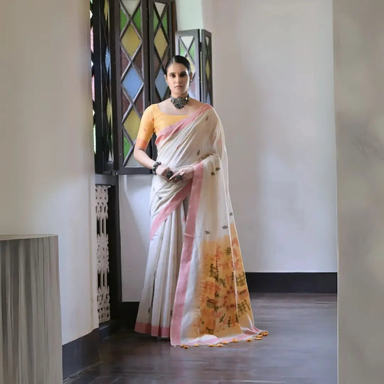 A woman models a graceful Off White Soft Muga Cotton Silk Saree. The saree has a pink accent border, and the pallu features a wide yellow panel decorated with traditional Warli-style figurative motifs. She wears a yellow blouse and a dark oxidized choker necklace, posing indoors.