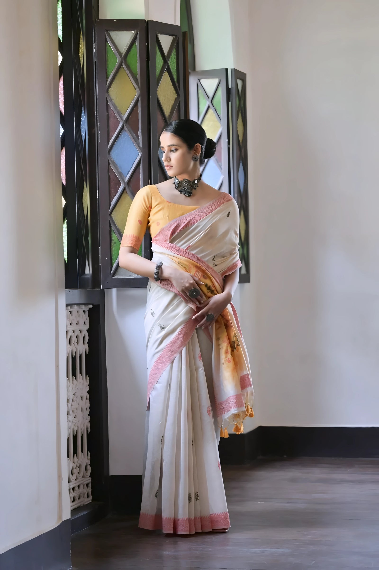 A woman models a graceful Off White Soft Muga Cotton Silk Saree. The saree has a pink accent border, and the pallu features a wide yellow panel decorated with traditional Warli-style figurative motifs. She wears a yellow blouse and a dark oxidized choker necklace, posing indoors.