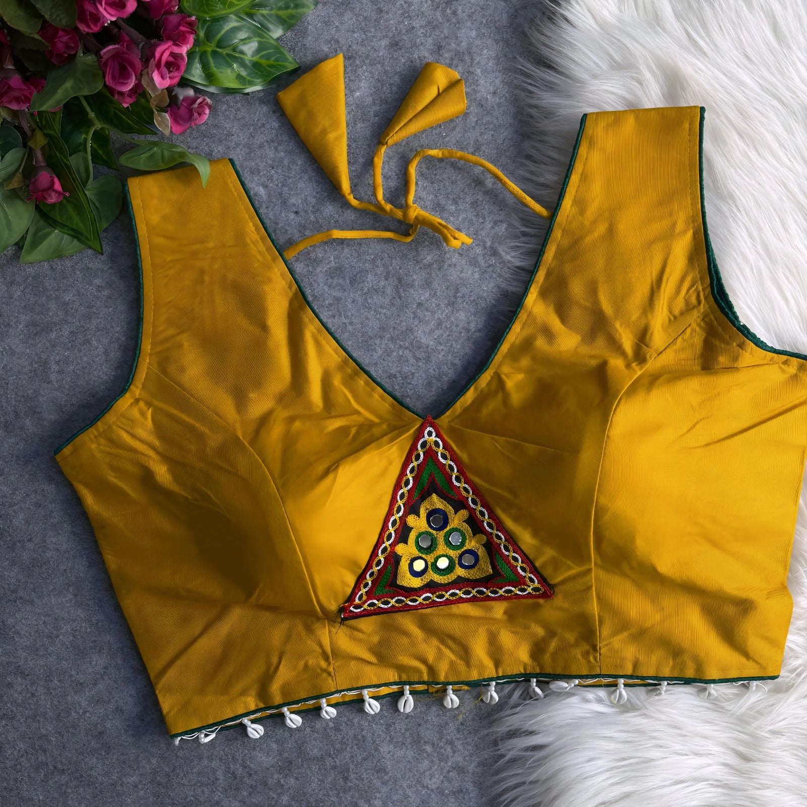 Yellow padded blouse with green piping, featuring a central embroidered triangle motif with mirror work and shell detailing along the hem, displayed on a grey surface with artificial pink flowers and white fur in the background.
