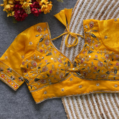 Yellow padded blouse with intricate multicolor floral embroidery, sequin accents, and a tie-back detail, displayed on a grey surface with artificial red and yellow flowers and a woven mat in the background.