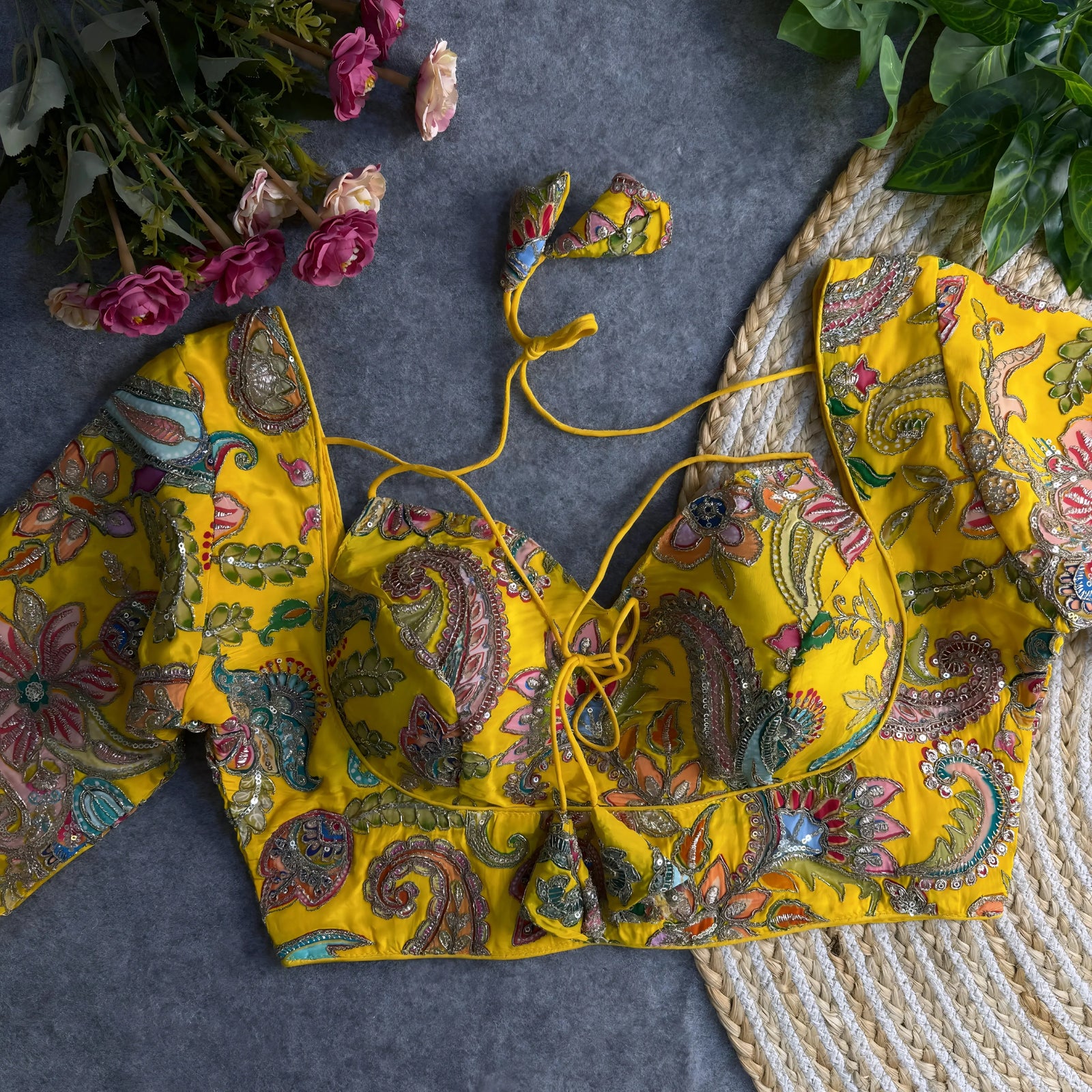 Yellow padded blouse with multicolor floral and paisley embroidery, featuring sequin accents and a tie-back detail, displayed on a grey surface with artificial pink flowers and green leaves in the background.