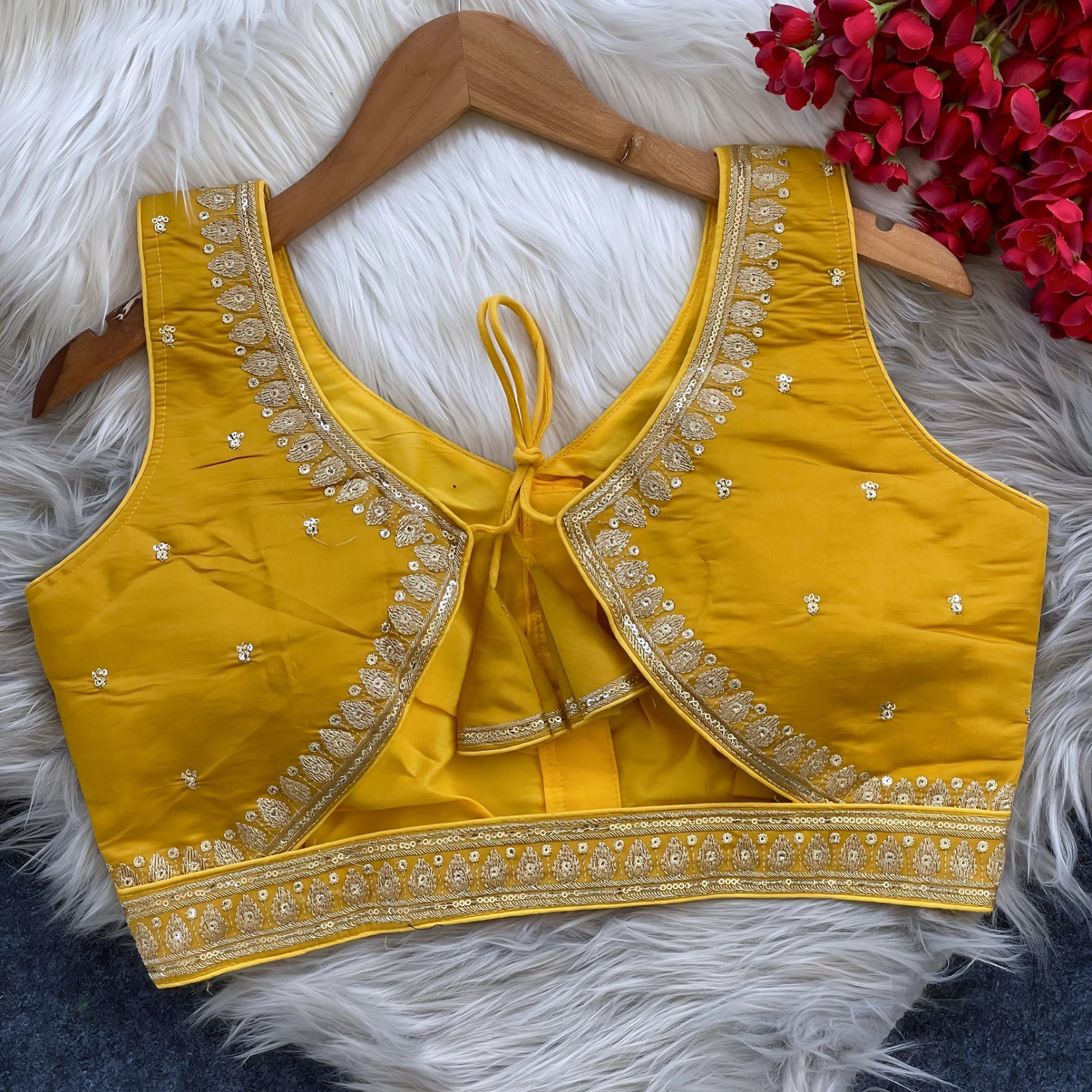 Yellow padded blouse with intricate gold embroidery along the neckline and hem, featuring small gold floral motifs, displayed on a wooden hanger over a white fur surface with red artificial flowers in the background.