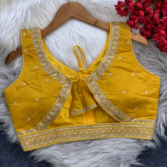 Yellow padded blouse with intricate gold embroidery along the neckline and hem, featuring small gold floral motifs, displayed on a wooden hanger over a white fur surface with red artificial flowers in the background.