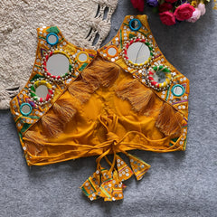 Traditional yellow Kutch-style blouse richly adorned with circular mirror work, multicolor rings, geometric thread embroidery, and decorative cowrie shells, featuring tasseled tie-back detail and displayed flat on a grey textured surface beside a braided cream mat and a bouquet of vibrant artificial roses.