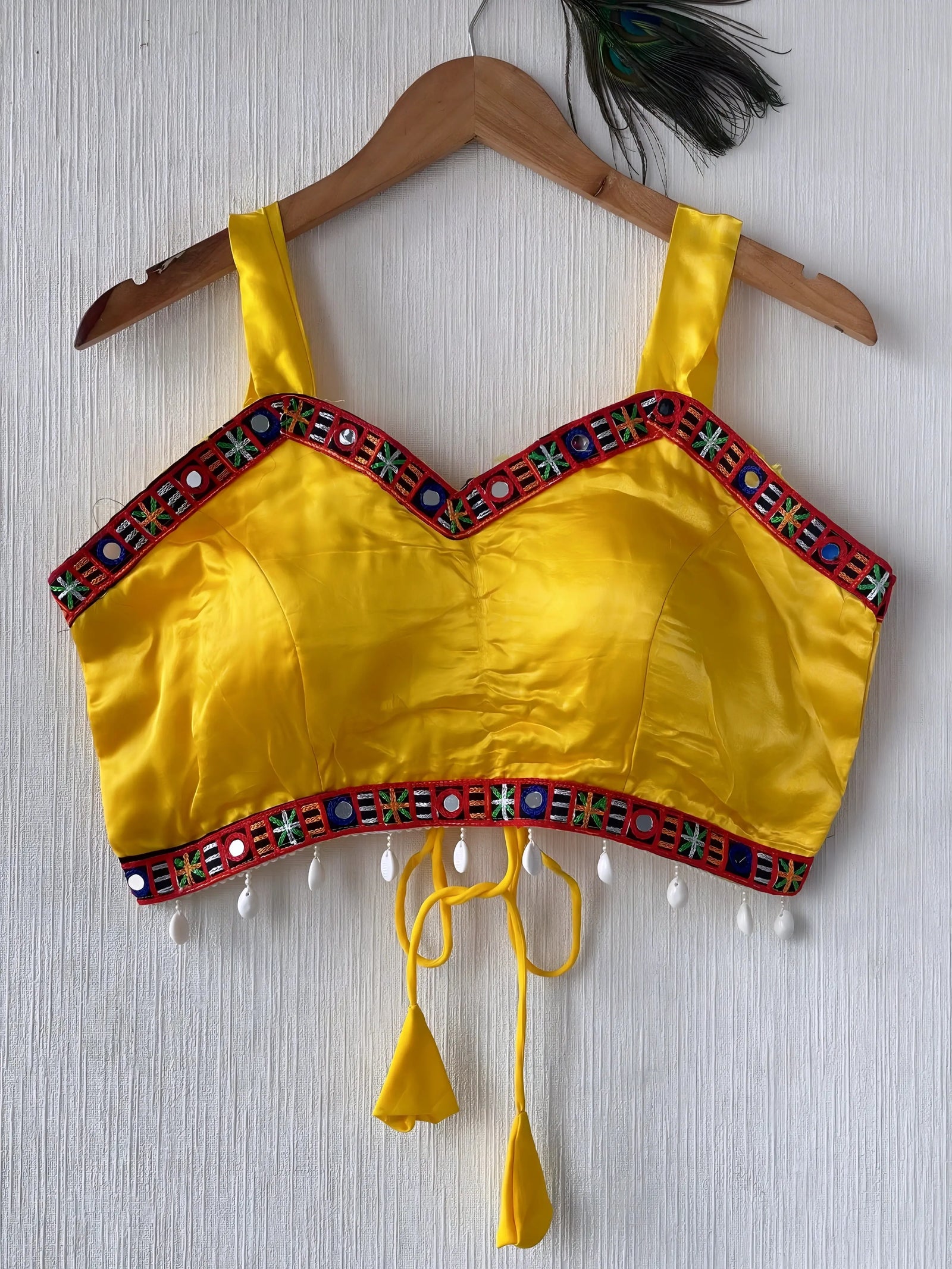 Bright yellow ethnic blouse featuring vibrant mirror work and embroidered border in red, green, and blue with pearl drop tassels and tie-up details, displayed flat on a hanger against a textured off-white wall for a festive and traditional look.
