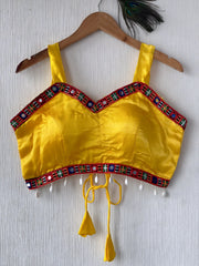 Bright yellow ethnic blouse featuring vibrant mirror work and embroidered border in red, green, and blue with pearl drop tassels and tie-up details, displayed flat on a hanger against a textured off-white wall for a festive and traditional look.