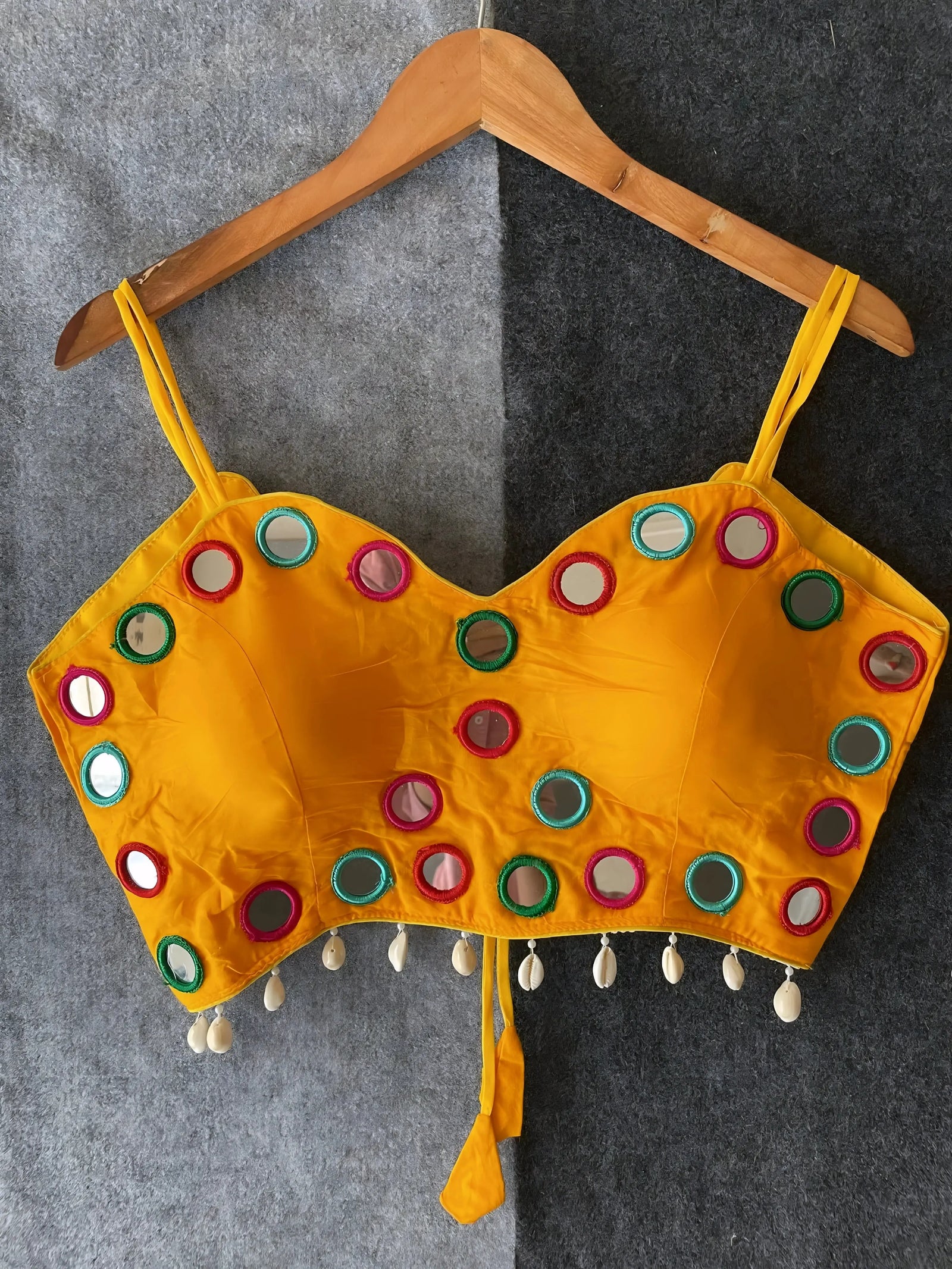Yellow corset-style blouse embellished with vibrant round mirrors and multicolored embroidered borders, featuring thin shoulder straps and shell tassel drops along the hem. Displayed on a wooden hanger against a two-tone grey textured background, showcasing its festive and contemporary ethnic design.
