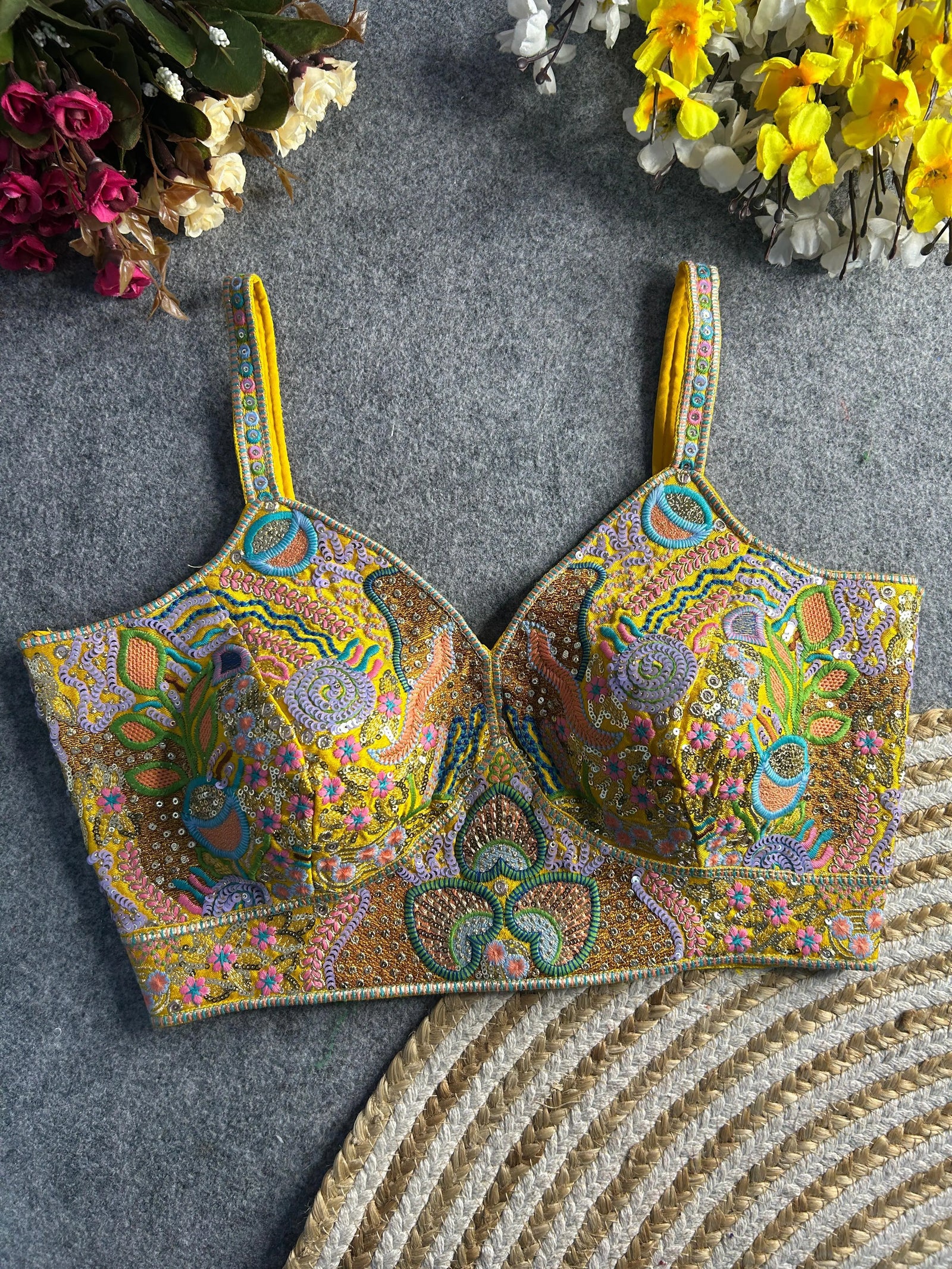 Traditional yellow bustier-style blouse featuring vibrant multicolor Madhubani-inspired embroidery with intricate floral, paisley, and geometric motifs, embellished with sequins and beads, structured cups, and adjustable straps, displayed flat on a textured grey surface with artificial pink, yellow, and white flowers in the corners.
Traditional yellow bustier-style blouse featuring vibrant multicolor Madhubani-inspired embroidery with intricate floral, paisley, and geometric motifs, embellished with sequin