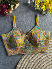 Traditional yellow bustier-style blouse featuring vibrant multicolor Madhubani-inspired embroidery with intricate floral, paisley, and geometric motifs, embellished with sequins and beads, structured cups, and adjustable straps, displayed flat on a textured grey surface with artificial pink, yellow, and white flowers in the corners.
Traditional yellow bustier-style blouse featuring vibrant multicolor Madhubani-inspired embroidery with intricate floral, paisley, and geometric motifs, embellished with sequin