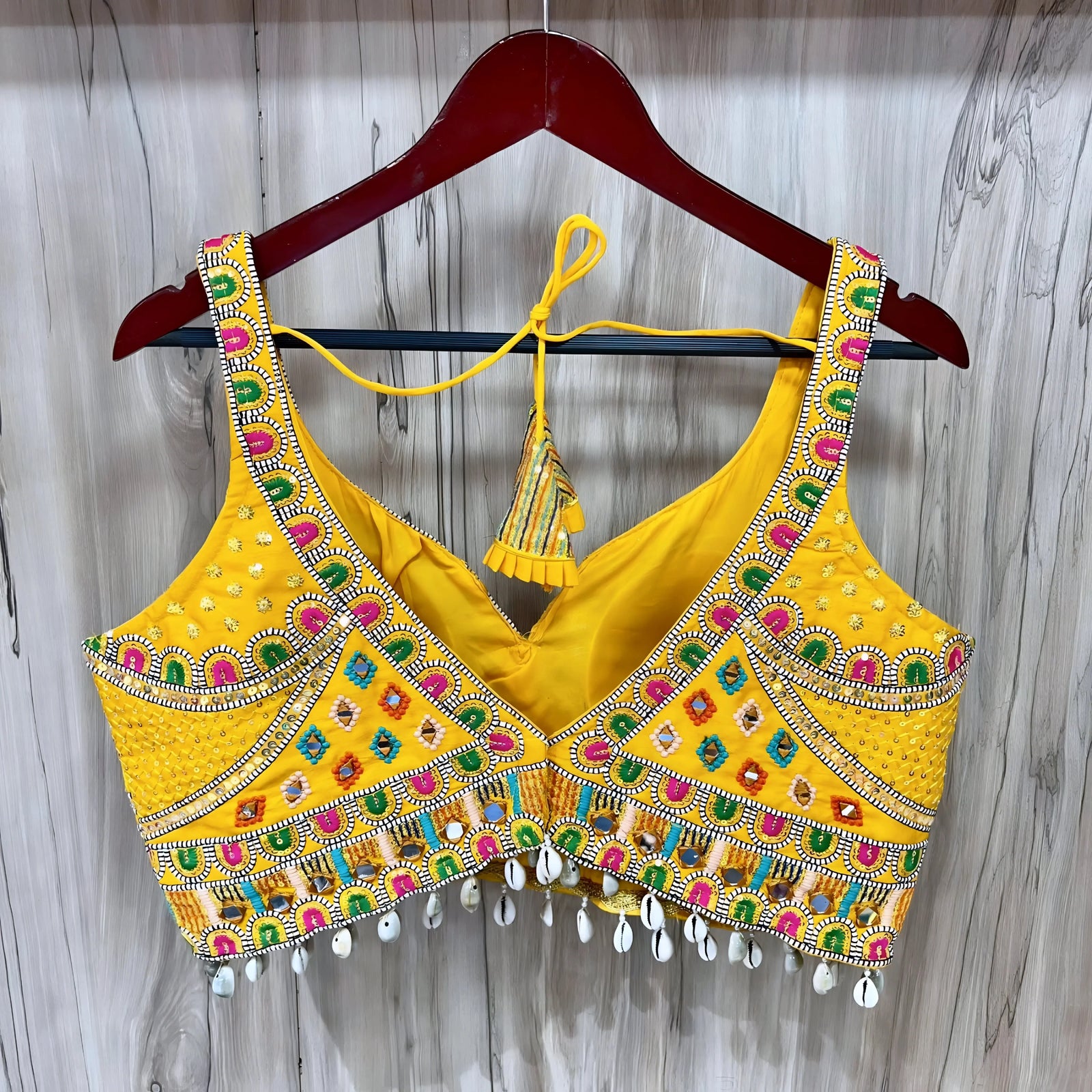 Traditional yellow Kutch-style blouse beautifully adorned with vibrant multicolor geometric embroidery, detailed mirror work including circular and teardrop shapes, accented with colorful beads and cowrie shell trim at the hem. The blouse features wide straps, structured cups, and tasseled yellow tie-back details, displayed flat on a grey textured surface with artificial pink flowers and green foliage at the corners.