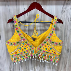Traditional yellow Kutch-style blouse beautifully adorned with vibrant multicolor geometric embroidery, detailed mirror work including circular and teardrop shapes, accented with colorful beads and cowrie shell trim at the hem. The blouse features wide straps, structured cups, and tasseled yellow tie-back details, displayed flat on a grey textured surface with artificial pink flowers and green foliage at the corners.