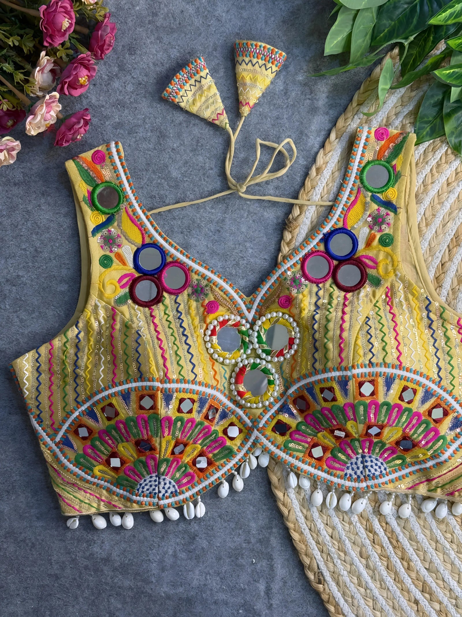 Traditional yellow blouse featuring vibrant multicolor mirror work, intricate thread embroidery in geometric and floral patterns, and shell tassel detailing along the hem, displayed on a flat surface with floral and leafy decor.