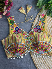 Traditional yellow blouse featuring vibrant multicolor mirror work, intricate thread embroidery in geometric and floral patterns, and shell tassel detailing along the hem, displayed on a flat surface with floral and leafy decor.