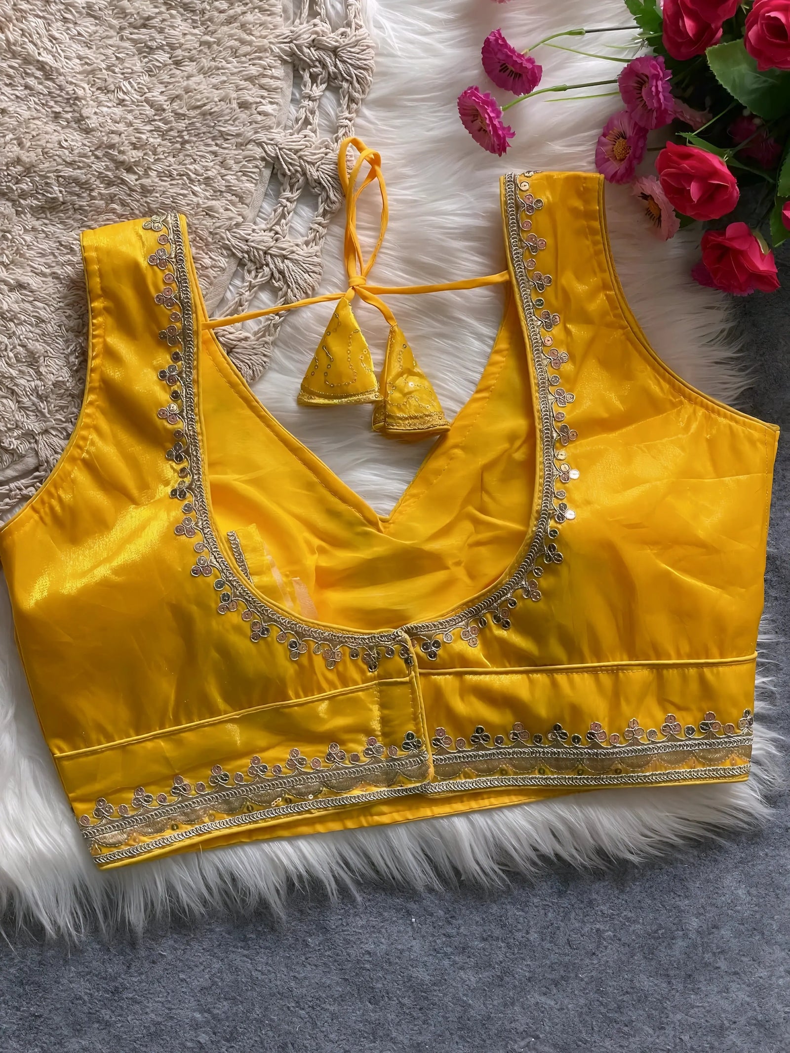 Traditional yellow silk blouse featuring intricate gold sequin and thread embroidery along the neckline and hem, with a decorative tassel tie at the back, displayed flat on a soft fur and textured mat background with artificial pink flowers in the corner.