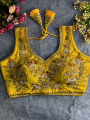 Traditional yellow silk blouse featuring intricate gold zardozi embroidery and beadwork with floral and leaf motifs on the front and neckline, finished with decorative tassel ties, displayed flat on a textured grey background with artificial pink and yellow flowers in the corners.