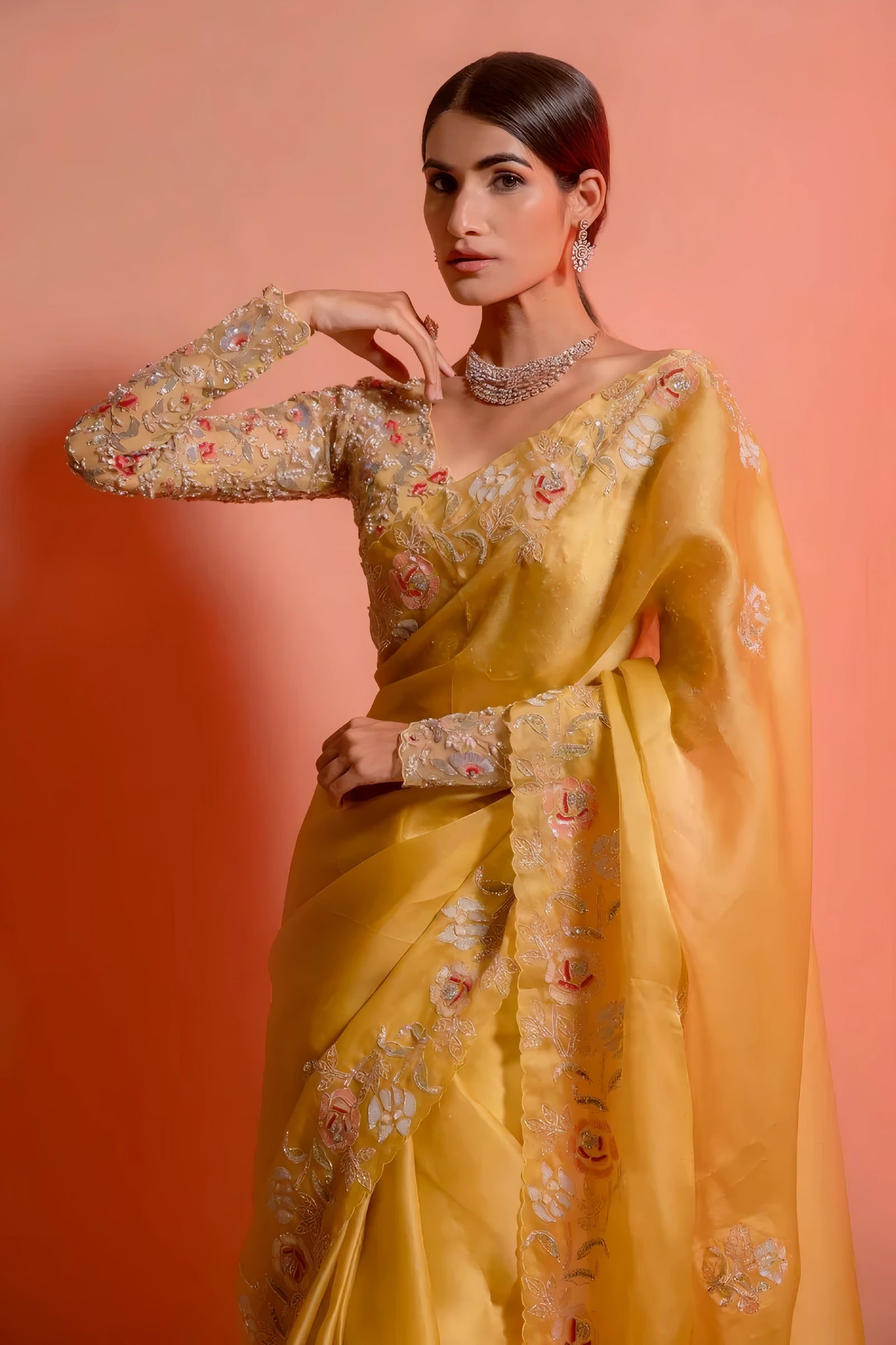 A woman models a striking Golden Yellow Soft Organza Silk Saree. The saree features delicate thread and sequin floral embroidery along the border. She wears a matching long-sleeve, heavily embroidered blouse and accessorizes with silver/diamond jewelry.