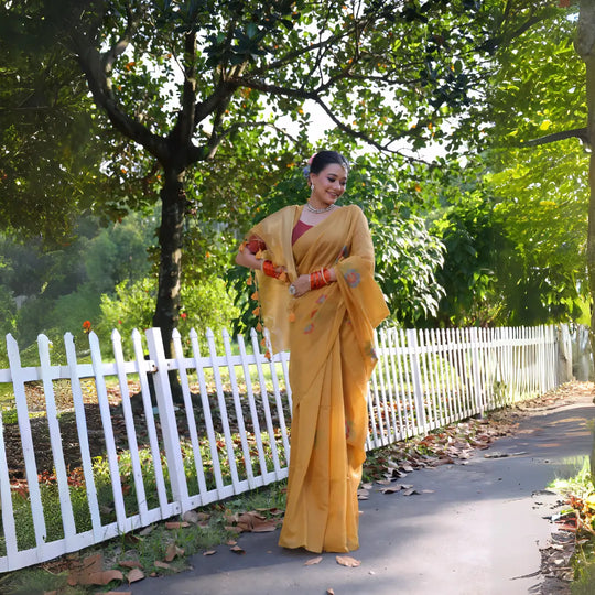 A woman models a vibrant Mustard Yellow Soft Muga Cotton Saree. The pallu features delicate colorful Resham floral weaving and tassels. She wears a maroon blouse and is accessorized with a white choker necklace and striking orange bangles, posing outdoors by a white fence.