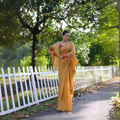 A woman models a vibrant Mustard Yellow Soft Muga Cotton Saree. The pallu features delicate colorful Resham floral weaving and tassels. She wears a maroon blouse and is accessorized with a white choker necklace and striking orange bangles, posing outdoors by a white fence.