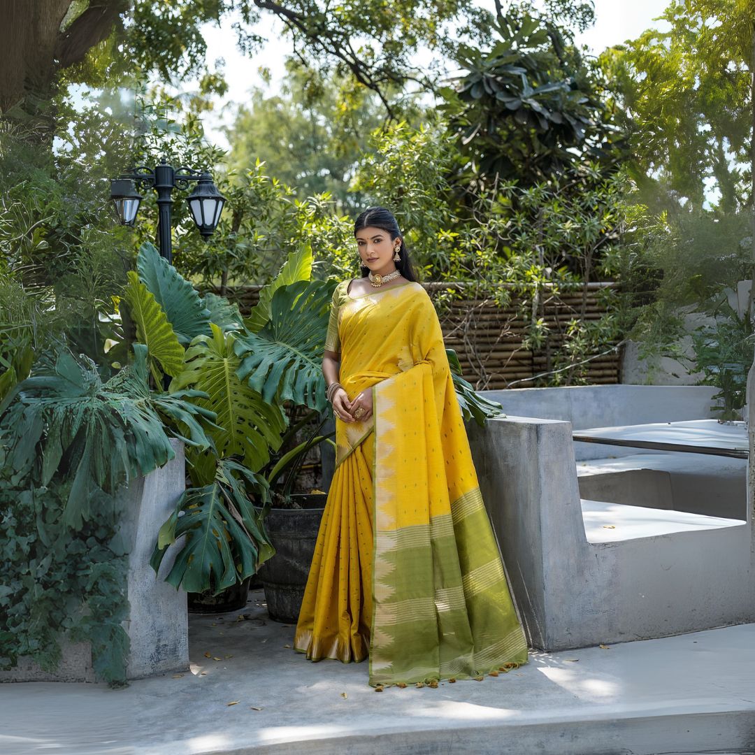 A woman models a vibrant Golden Yellow Tussar Silk Saree with small woven motifs. The pallu features wide bands of contrasting olive green and horizontal gold Zari stripes. She wears a light green blouse and heavy gold Kundan jewelry, posing outdoors.