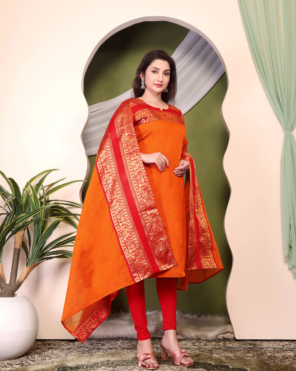 Woman wearing an orange and red traditional outfit with a plant and decorative archway in the background