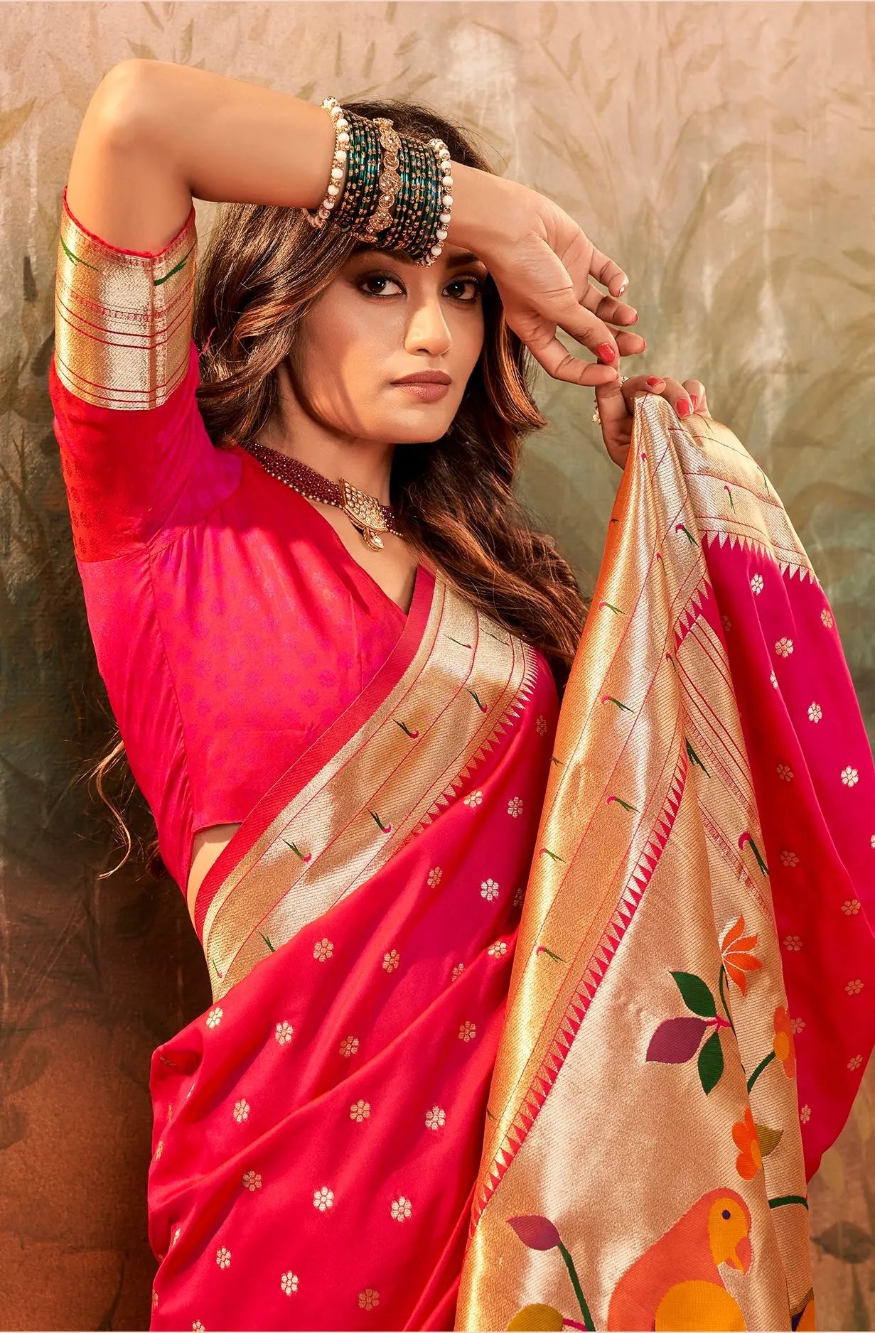 Woman in a red saree with gold border holding a matching saree against a textured wall.