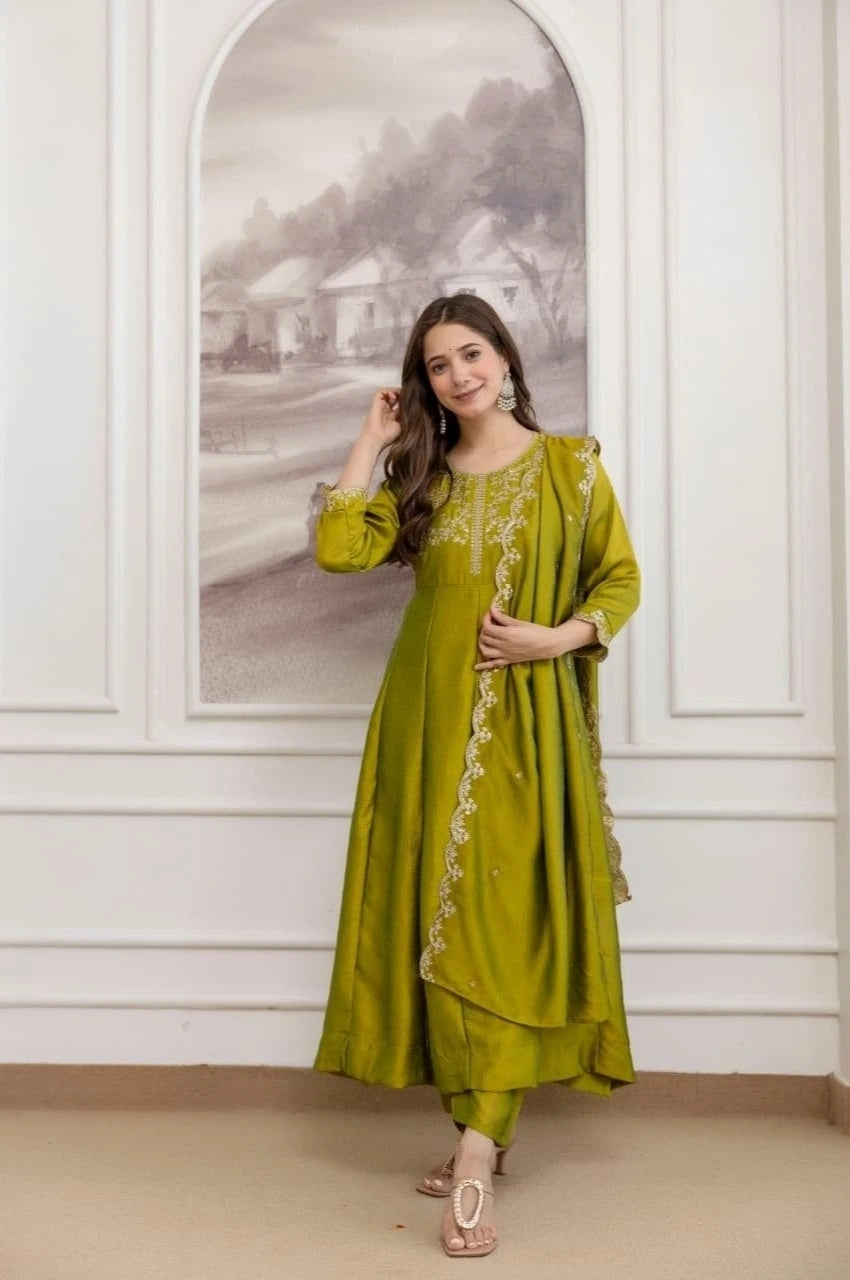 Woman wearing a green traditional outfit with intricate designs in a room with white walls and a framed picture.