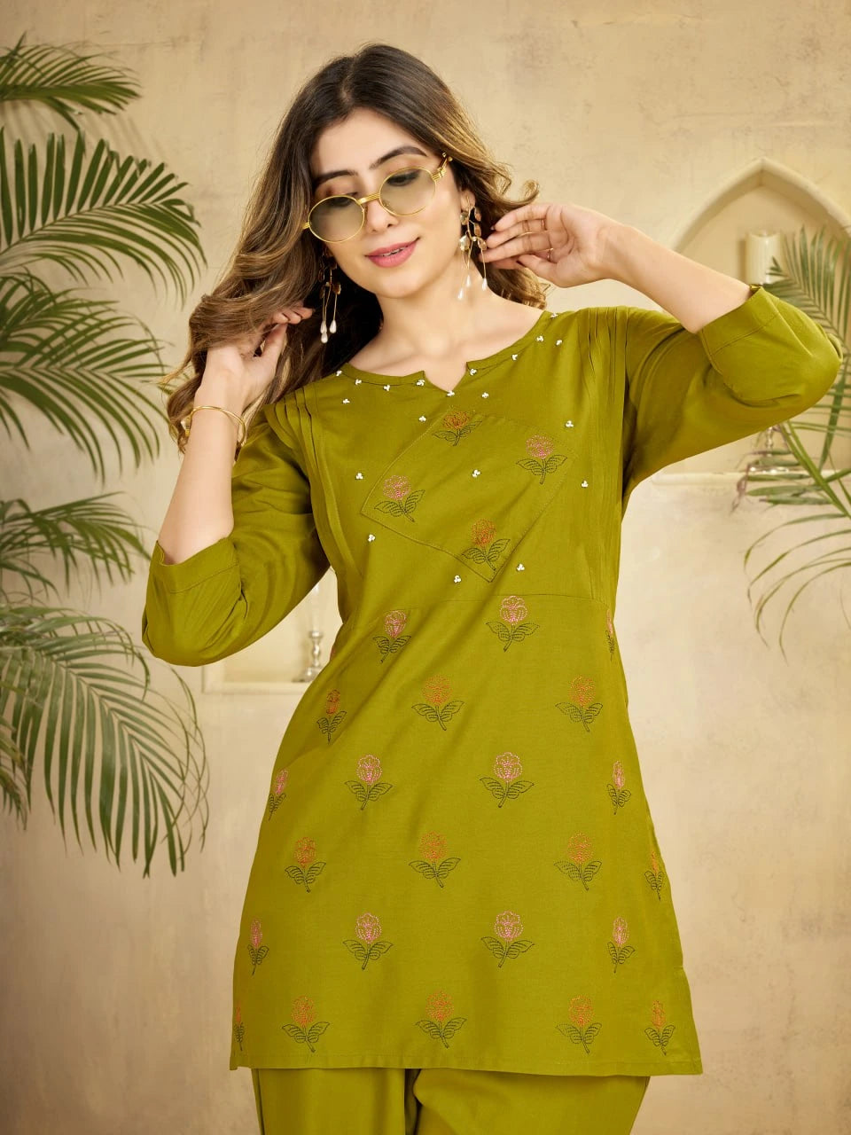 Woman wearing a green traditional outfit with floral patterns against a beige wall.