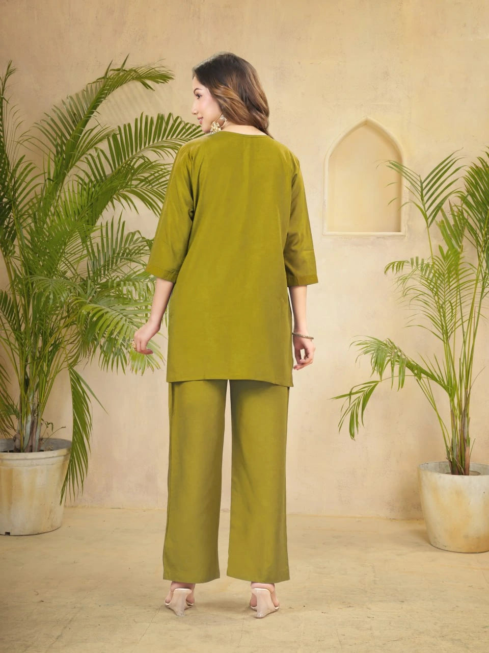 Woman in a green outfit standing between two potted plants in a room with beige walls.