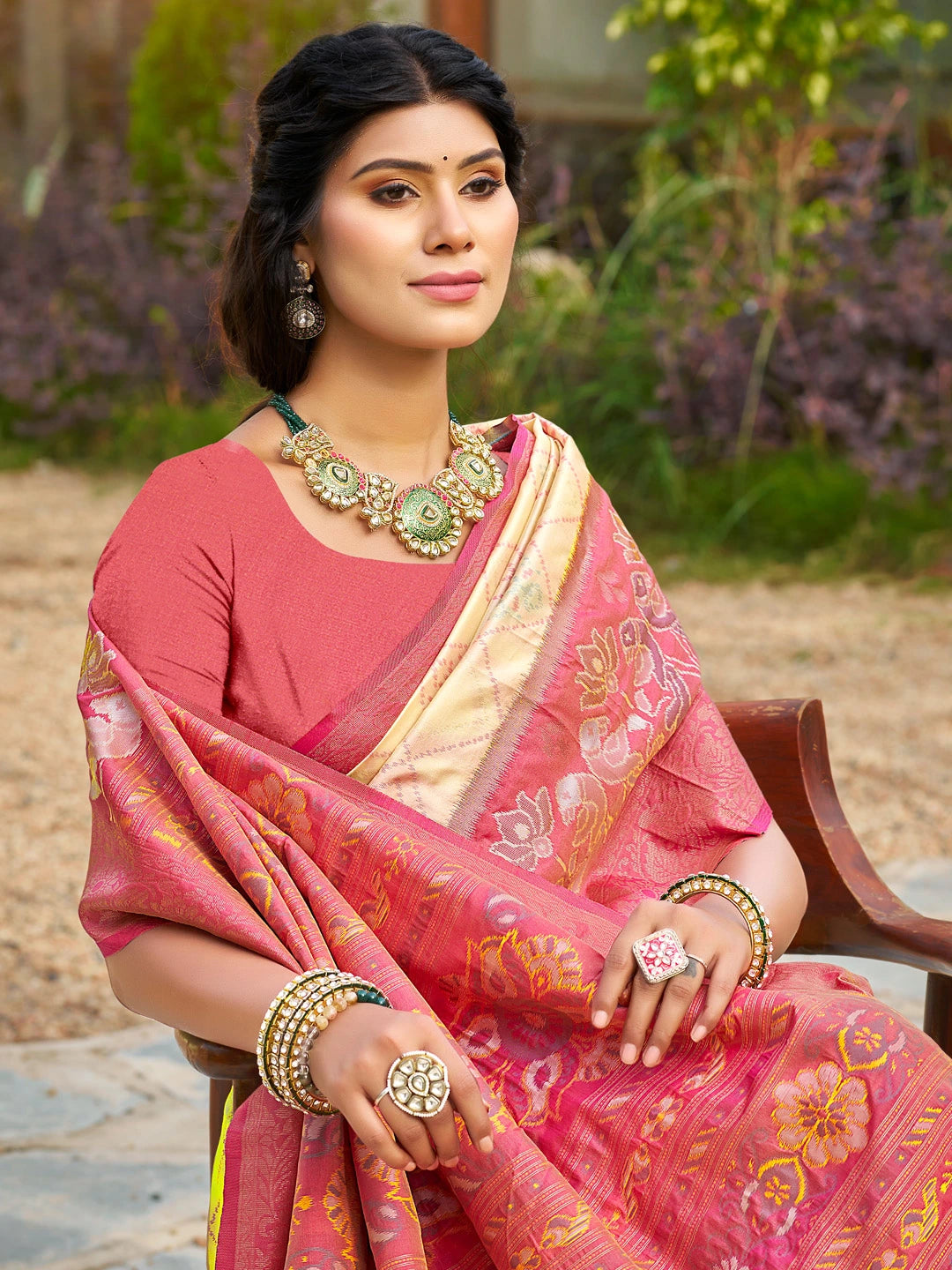 Woman in a pink saree with gold jewelry sitting outdoors.