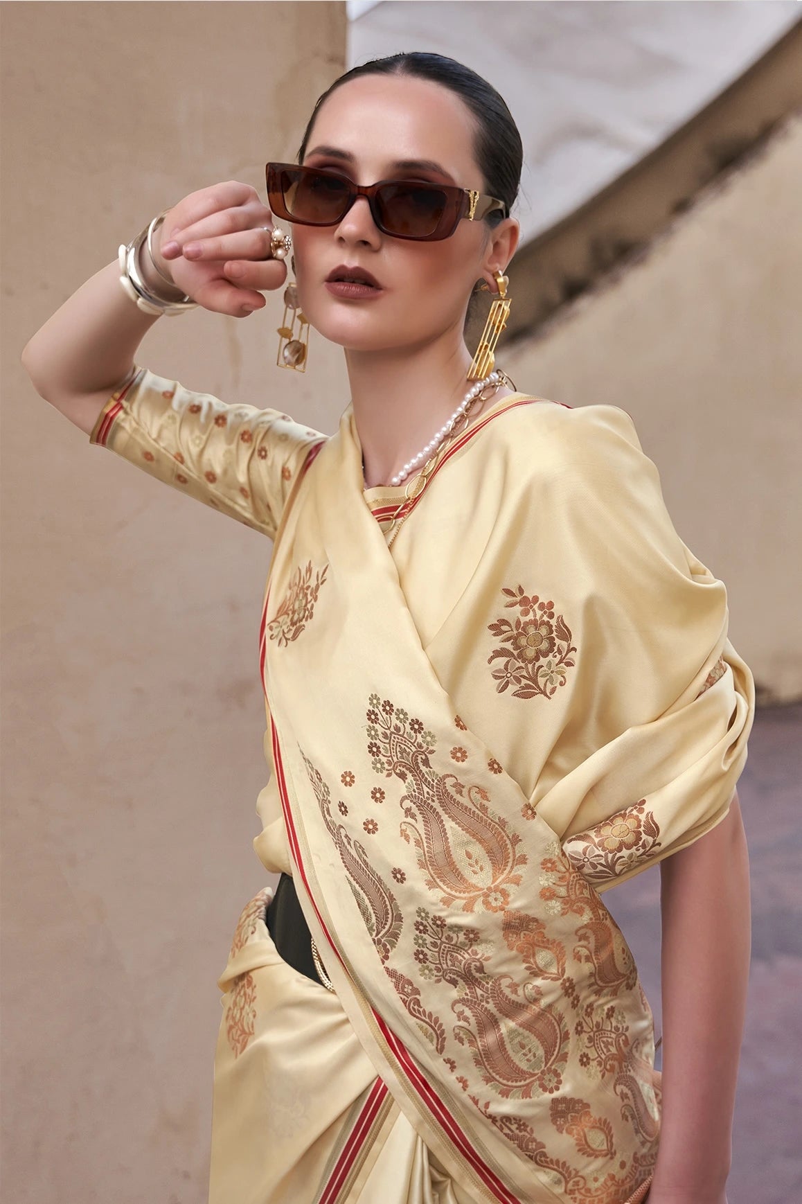 Woman wearing a cream saree with gold patterns and sunglasses against a neutral background