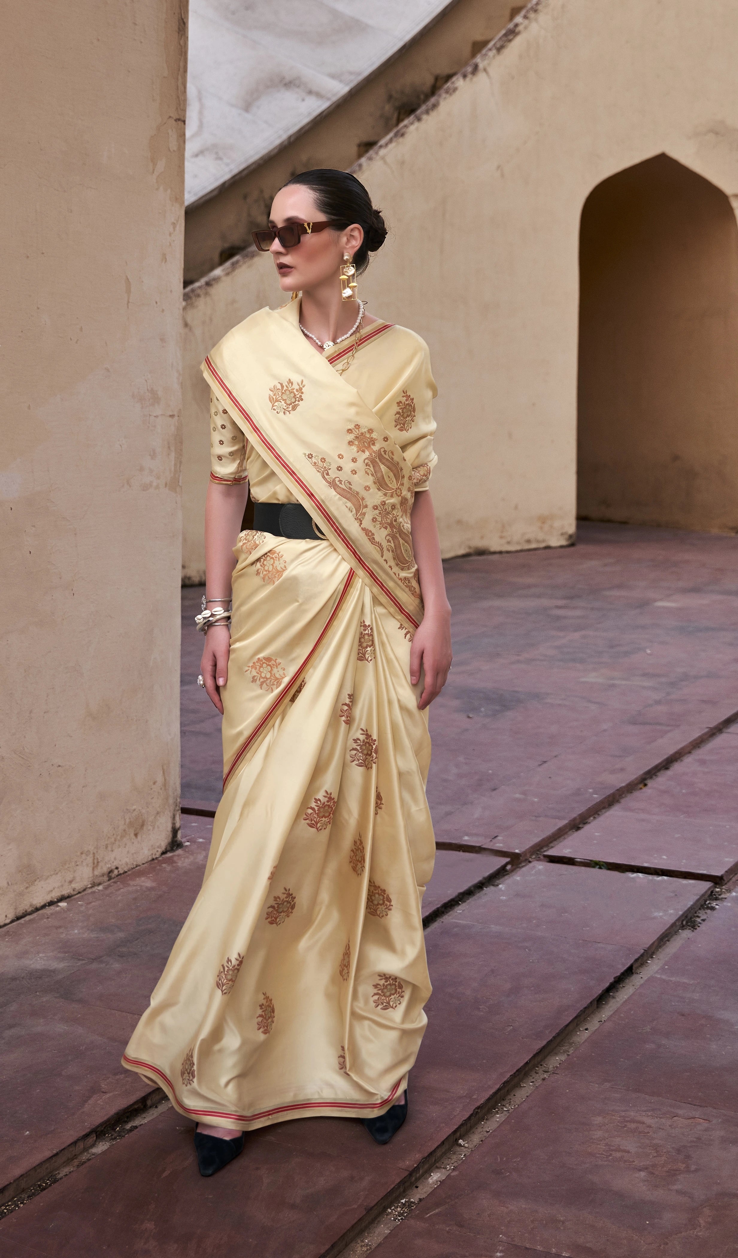 Woman in a cream saree with gold patterns standing in an architectural setting.