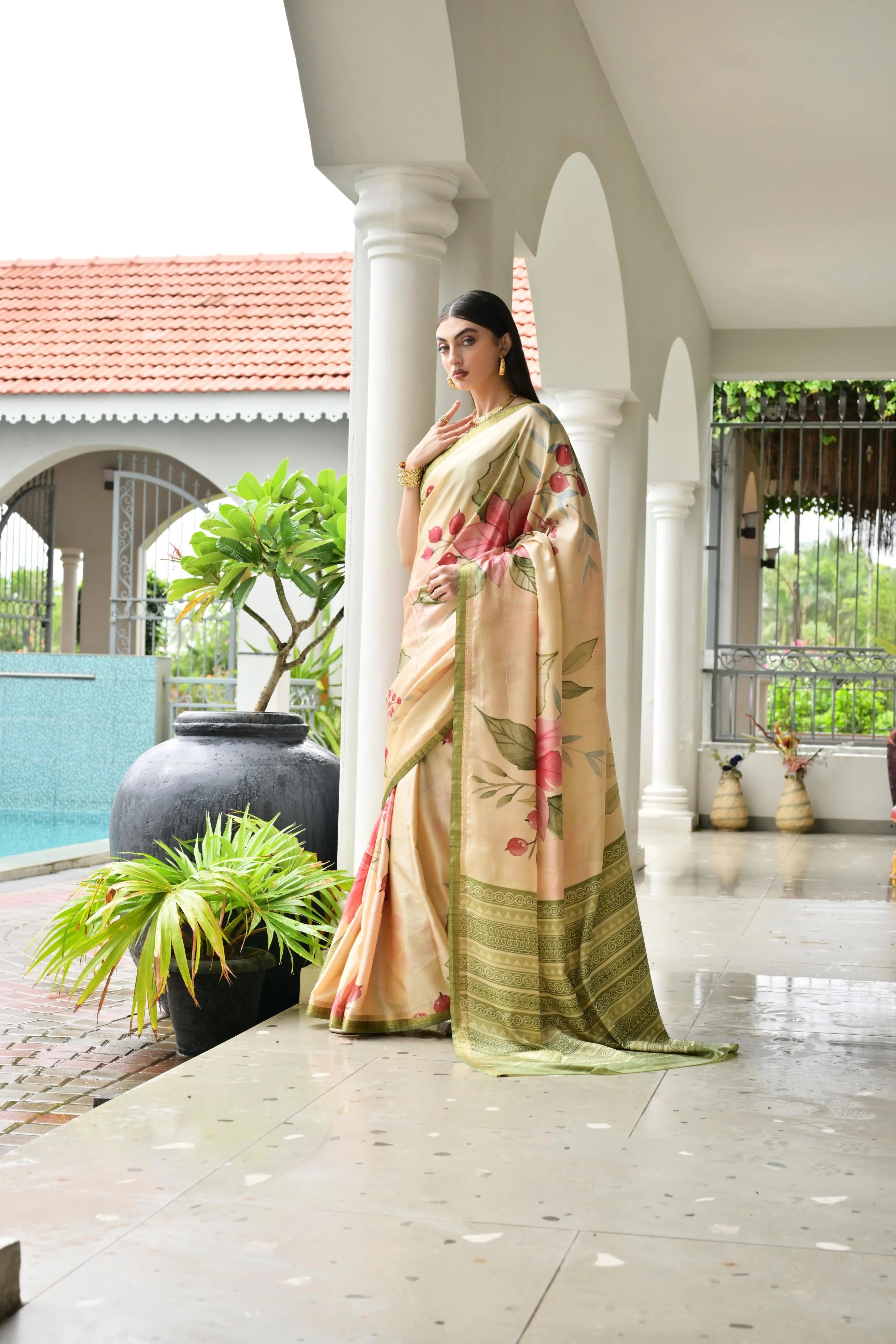 Woman in a traditional saree standing in a well-lit outdoor setting with plants and a pool.