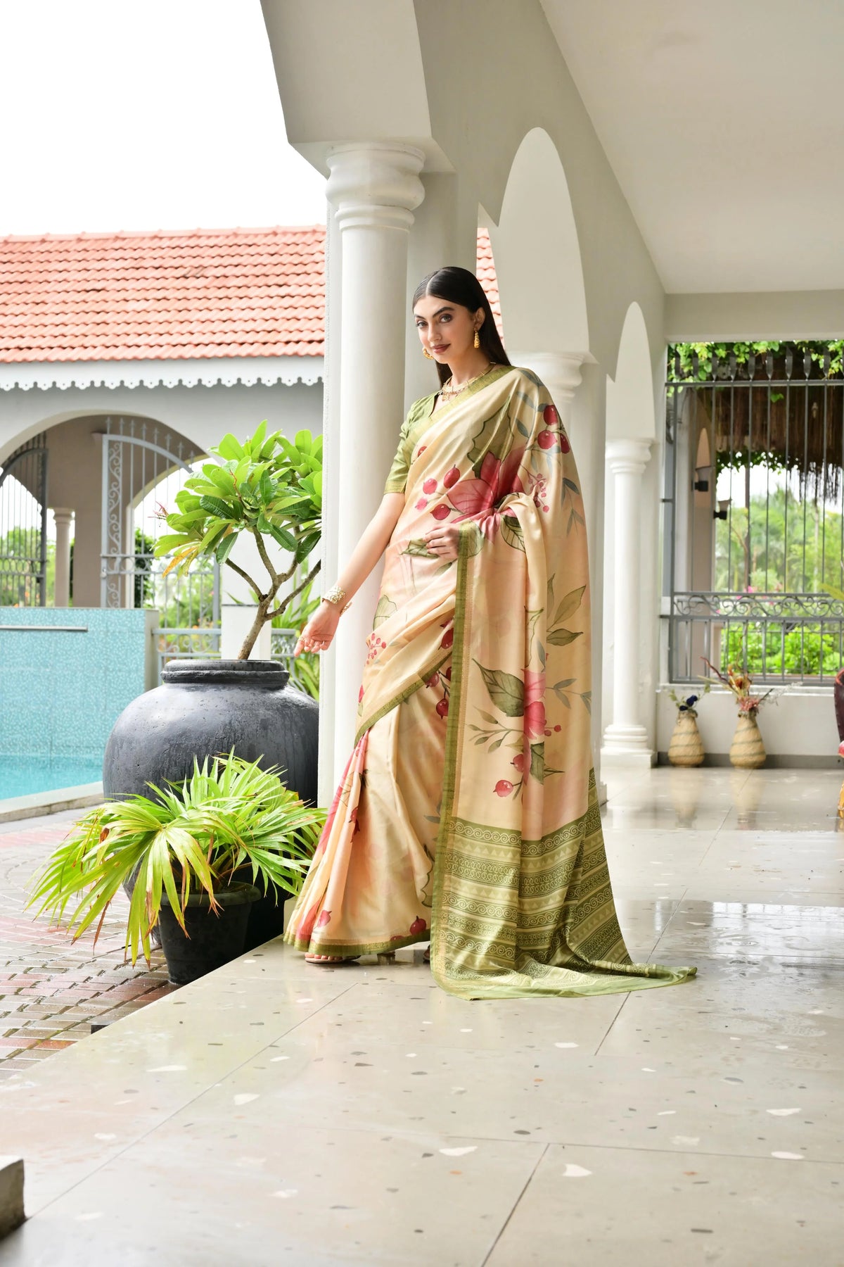 Woman in a traditional saree standing in a well-lit outdoor setting with plants and a pool.