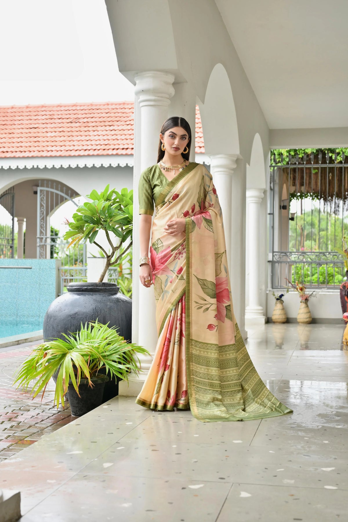Woman in a traditional saree standing in a decorative outdoor setting with plants and architectural elements.