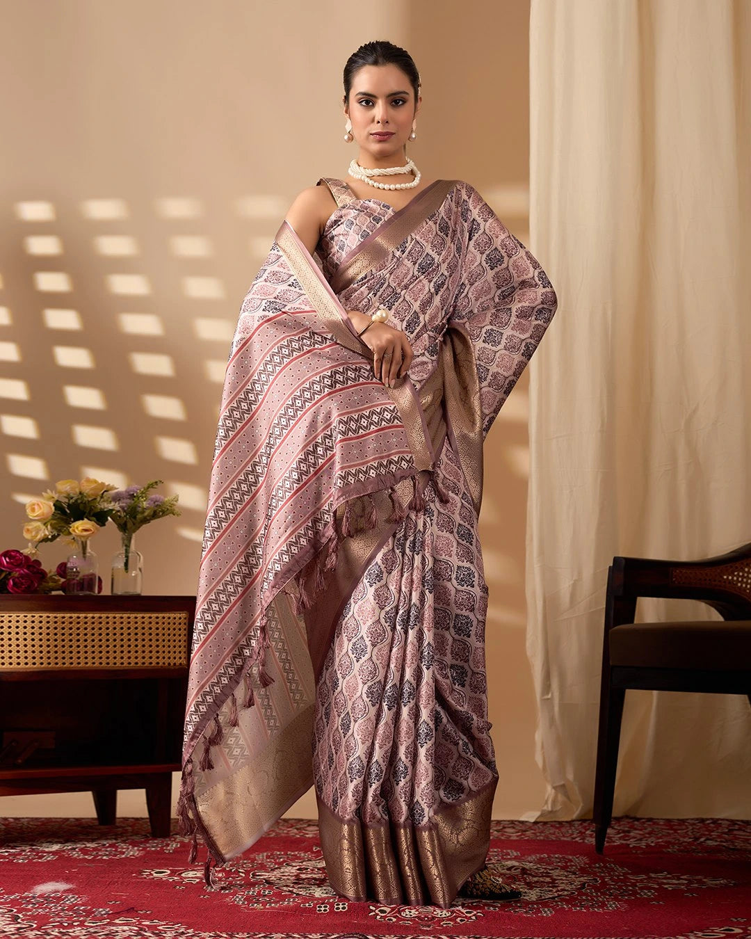 Woman wearing a patterned saree in a room with a table and flowers.