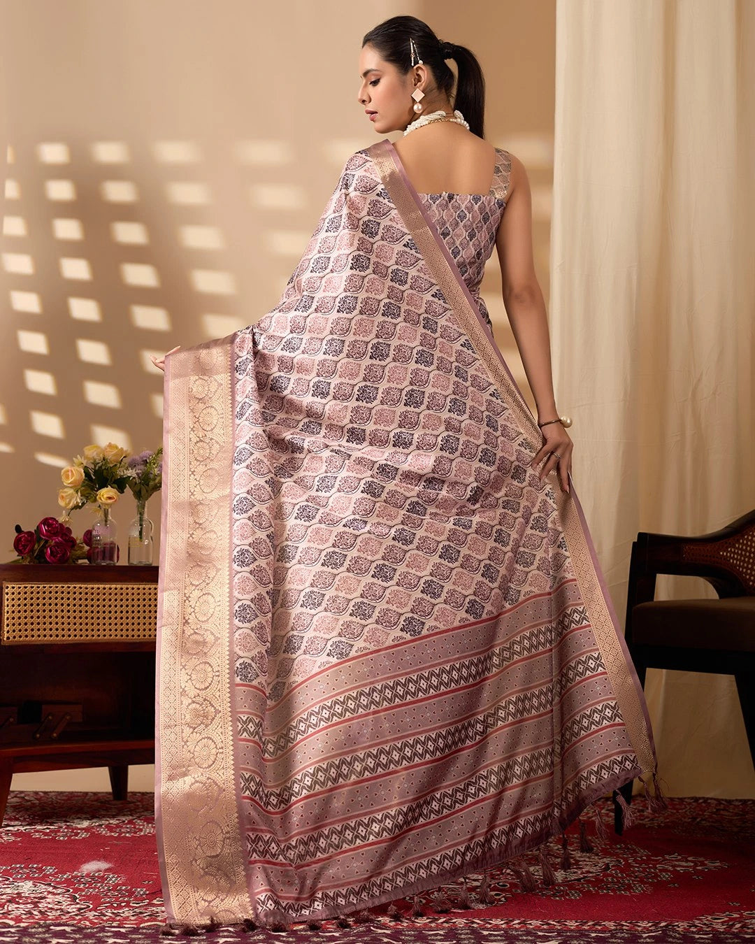 Woman wearing a patterned saree in a room with a table and flowers.