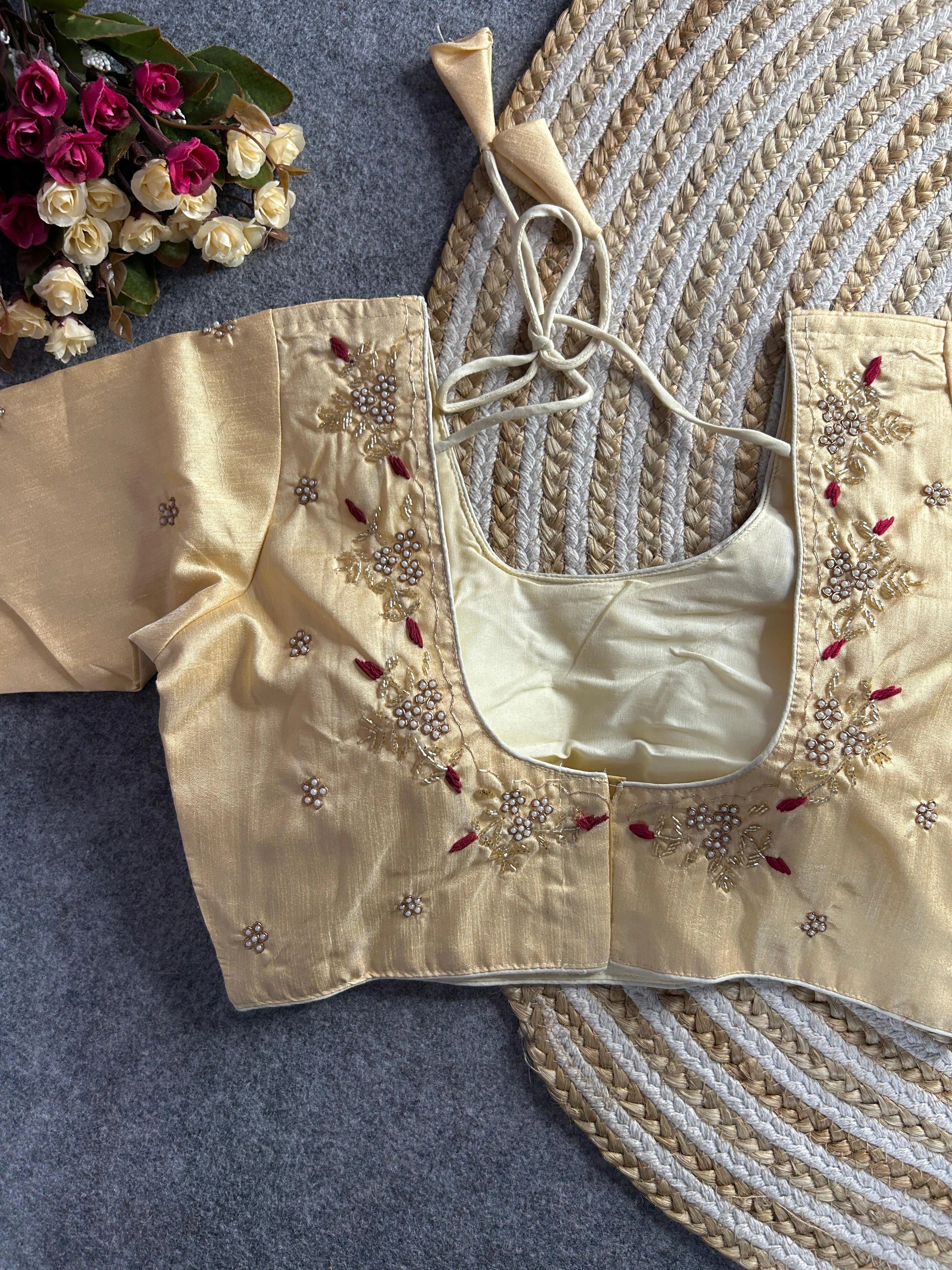 A soft cream-colored raw silk blouse with a wide U-neckline at the back. The neckline is bordered with delicate gold and pearl floral embroidery, featuring deep maroon threadwork accents. It includes matching cream tie-back cords with triangular tassels, and small pearl buttis are scattered across the fabric.