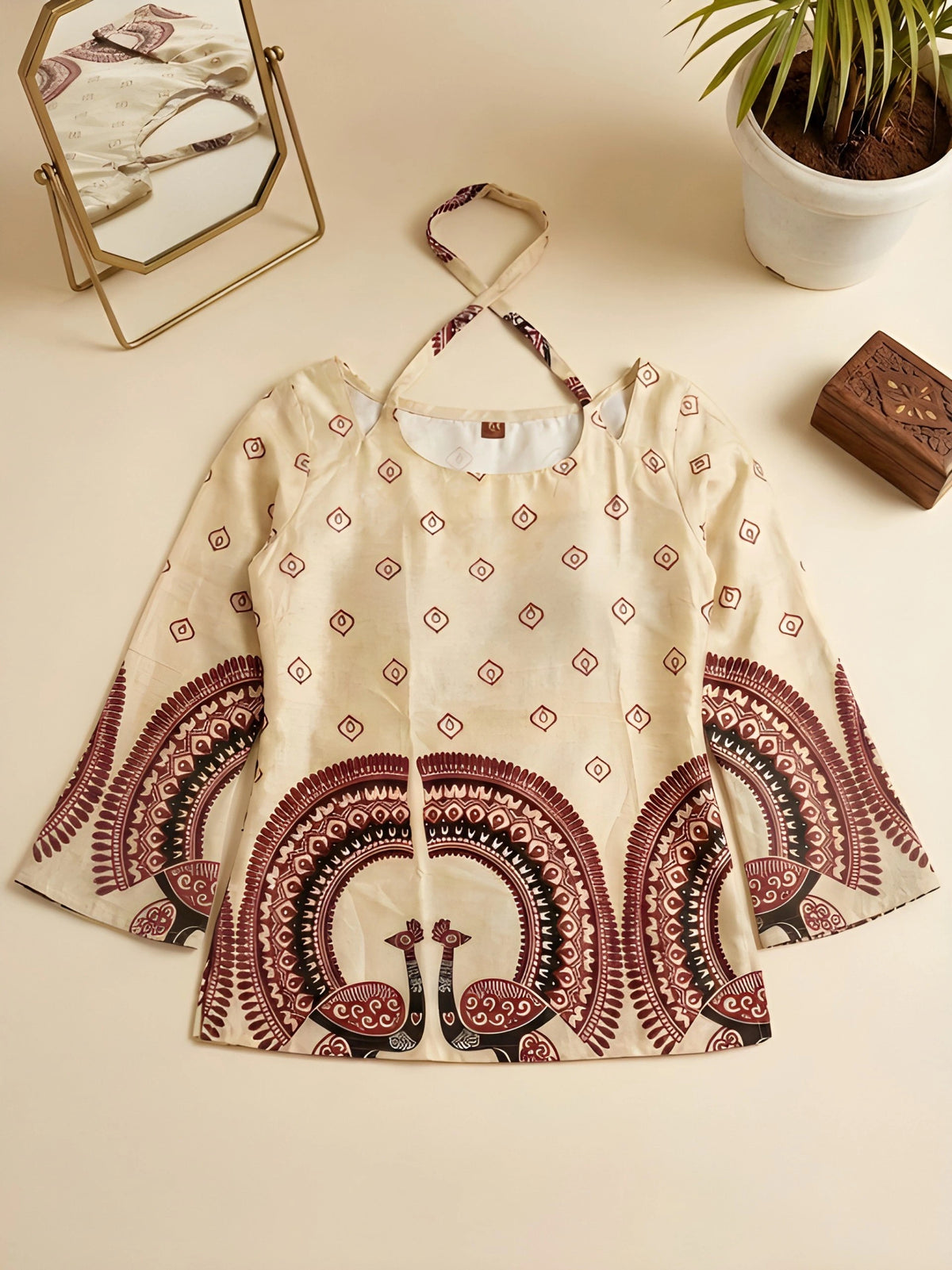 Beige blouse with red and black peacock pattern on a beige surface with a plant and mirror in the background.
