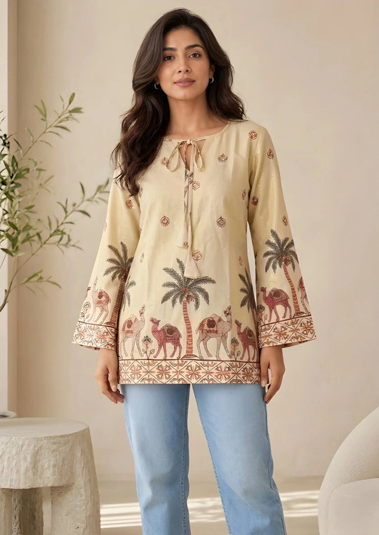 Woman wearing a beige embroidered top with palm tree and camel designs, standing indoors.