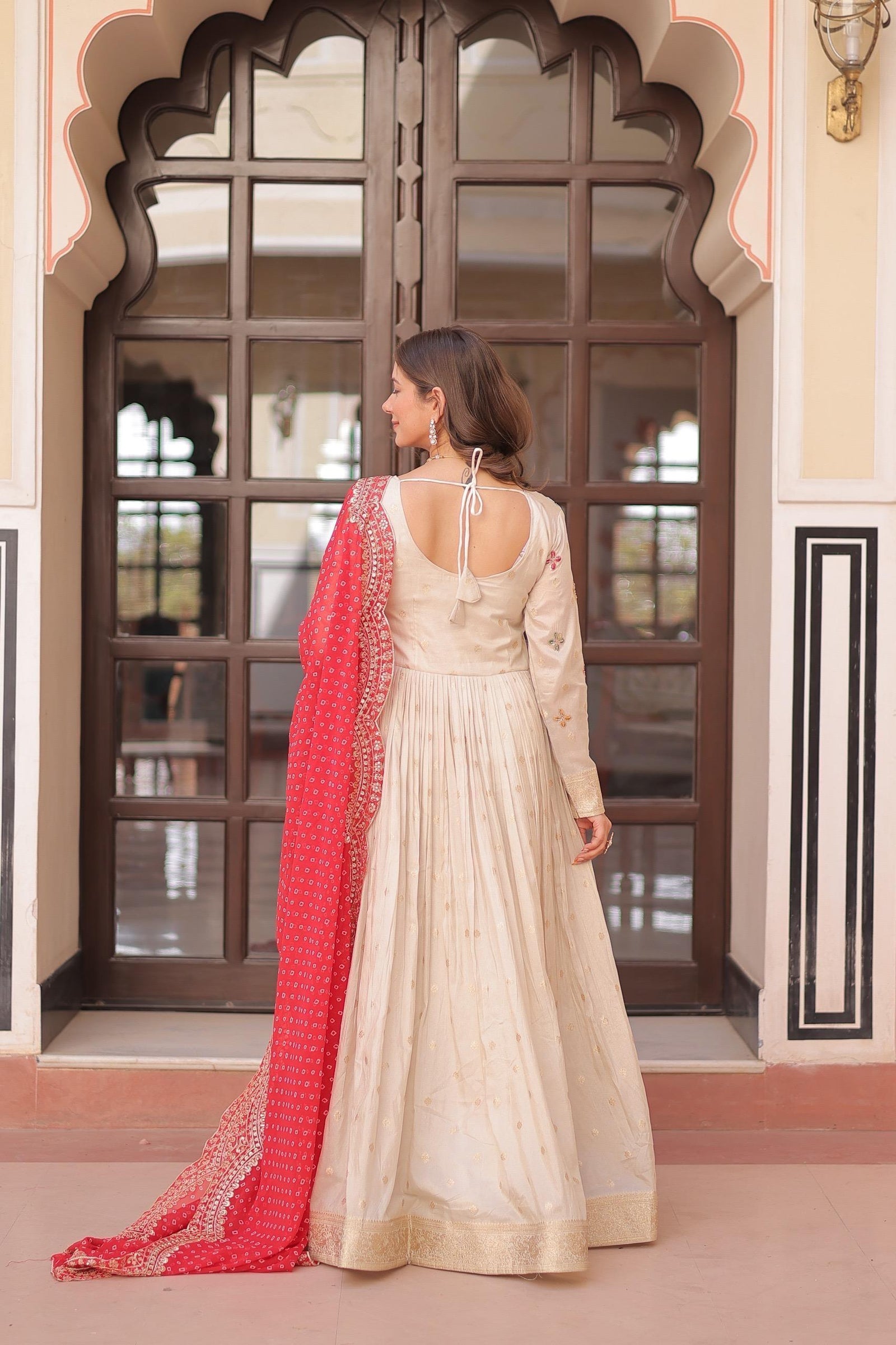 Woman in a cream traditional outfit with a red dupatta standing in front of an ornate doorway.