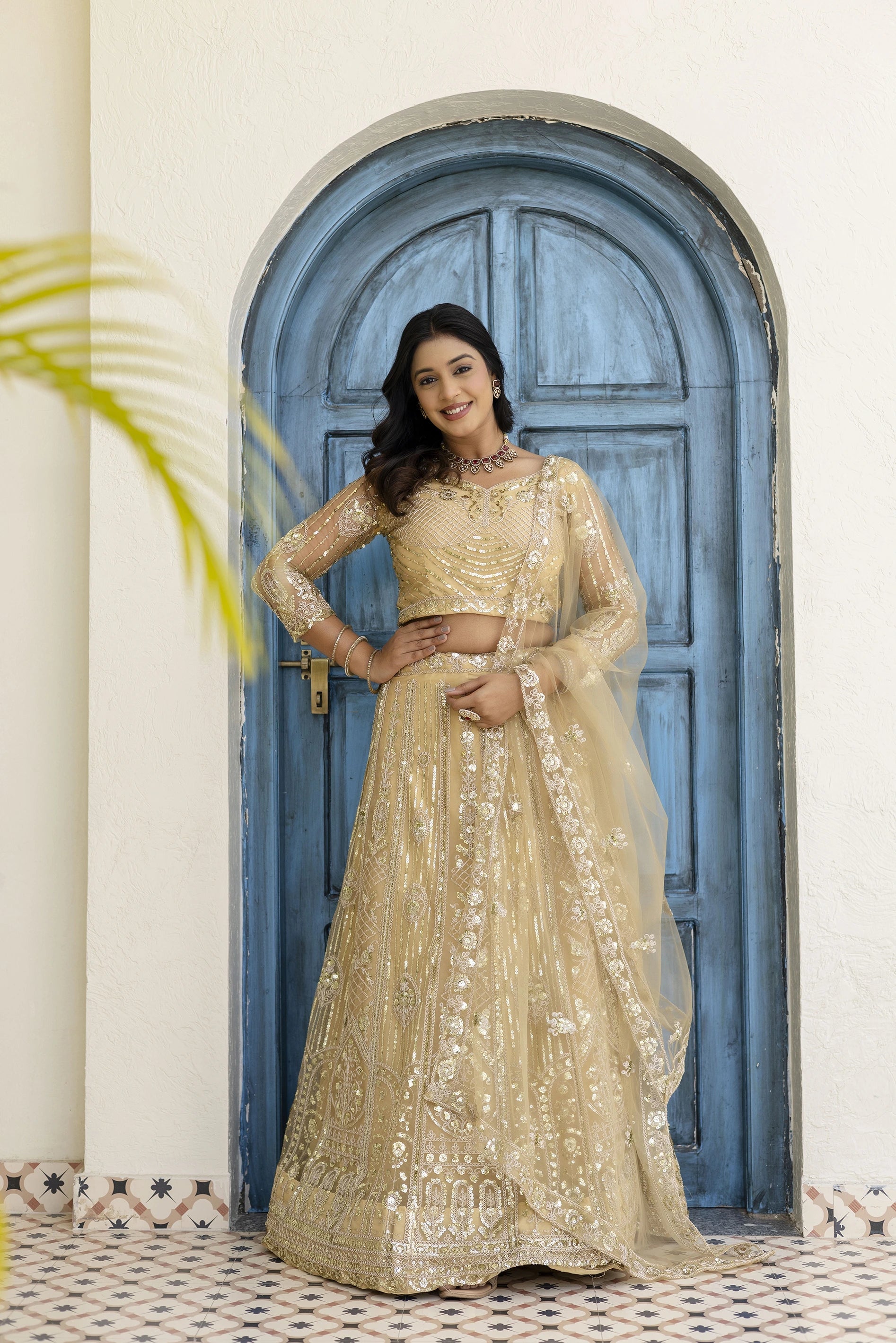 Woman in a beige embroidered lehenga standing in front of a blue door.