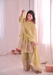 Woman in a light green traditional outfit with gold embroidery standing against a white curtain.