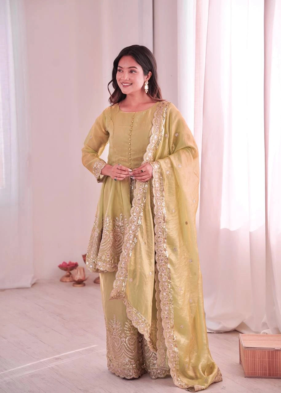 Woman in a light green traditional outfit with gold embroidery standing in a softly lit room.