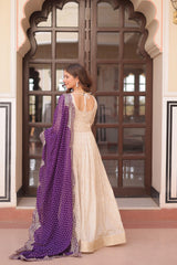 Woman in a white saree with a purple shawl standing in front of an ornate door.