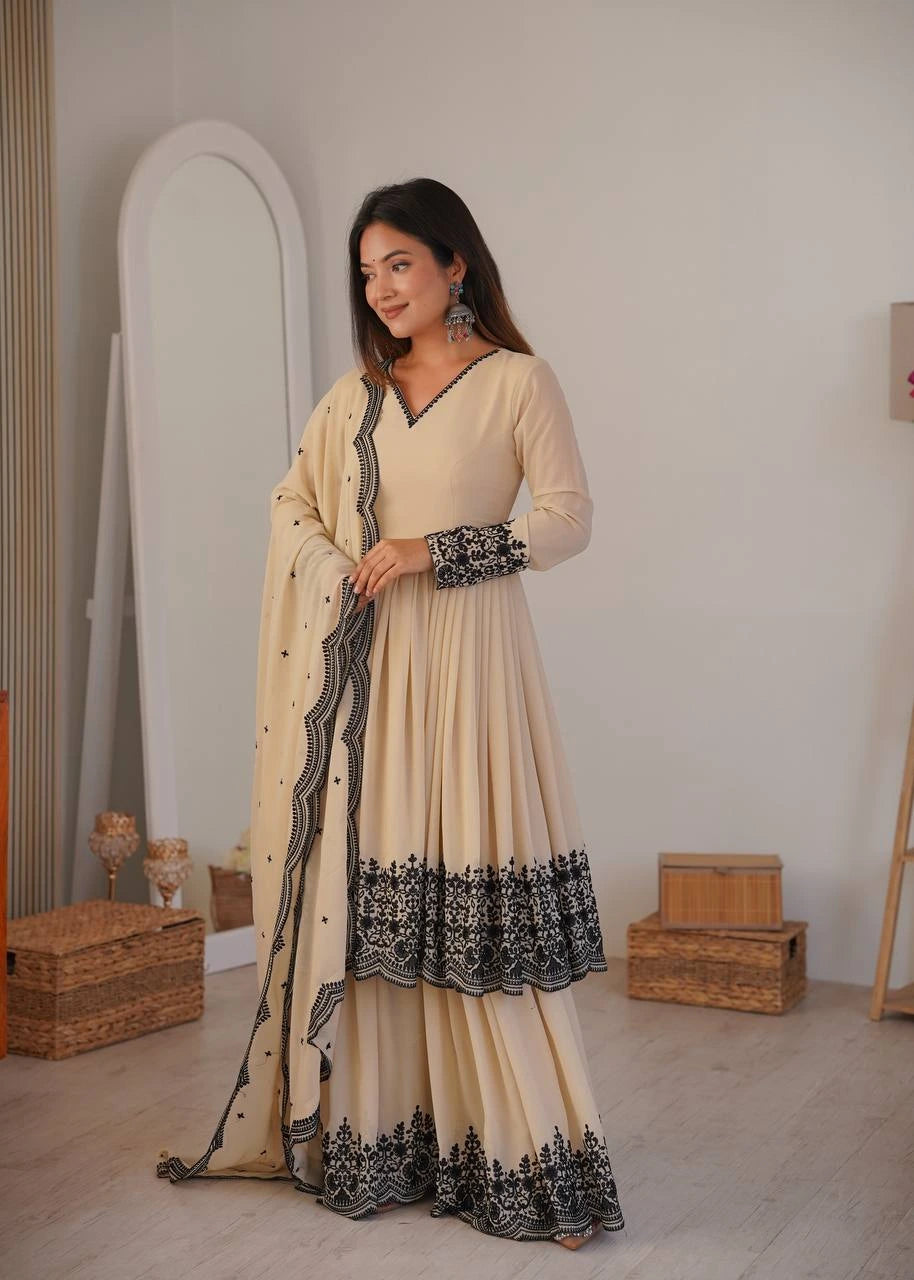 Woman in a beige and black traditional outfit standing in a room with wooden boxes and a mirror.