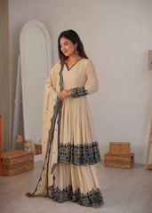 Woman in a beige and black traditional outfit standing in a room with wooden boxes and a mirror.