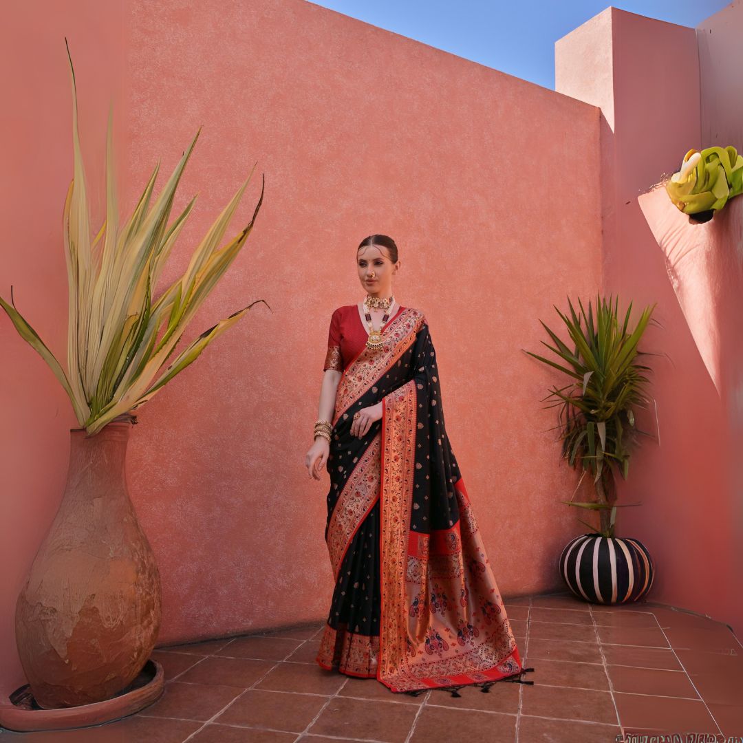 A woman models a striking black and red Banarasi Silk Saree. The saree is black with intricate Zari weaving across the body and features a wide, ornate red/gold Zari woven border and pallu. She pairs it with a short-sleeve red blouse and heavy traditional gold jewelry.