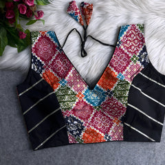 Traditional multicolor Ajrak-print blouse featuring vibrant geometric and floral motifs in orange, pink, blue, green, and red. The blouse has a deep V-neckline, black silk side panels with metallic silver stripes, and a patterned tasseled tie-back, displayed flat on a white fur background with artificial pink flowers on the side.